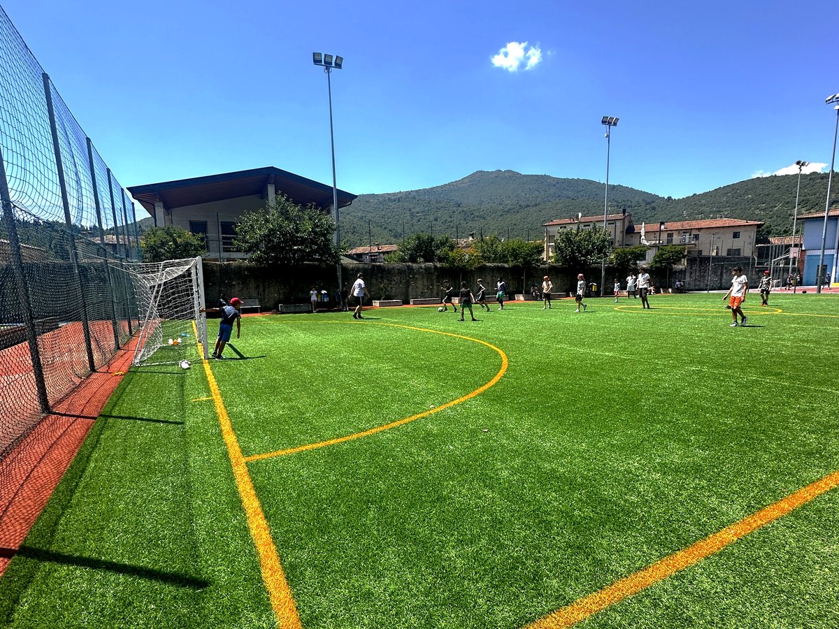 Campo da calcio in erba sintetica con partita in corso e vista montagne